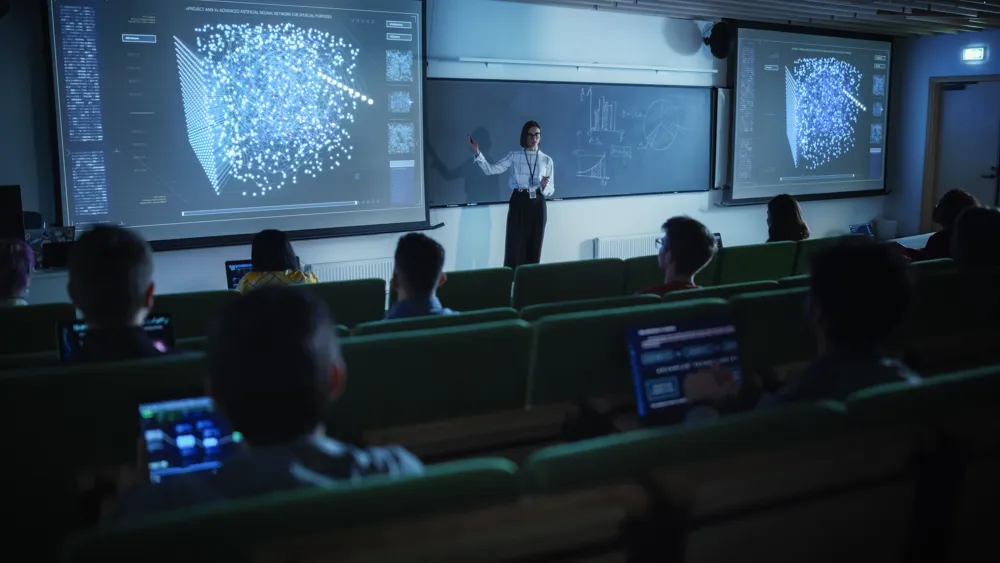 ALT text: A female presenter in a tech-focused classroom explains a 3D quantum displayed on two large screens, addressing students seated with laptops.